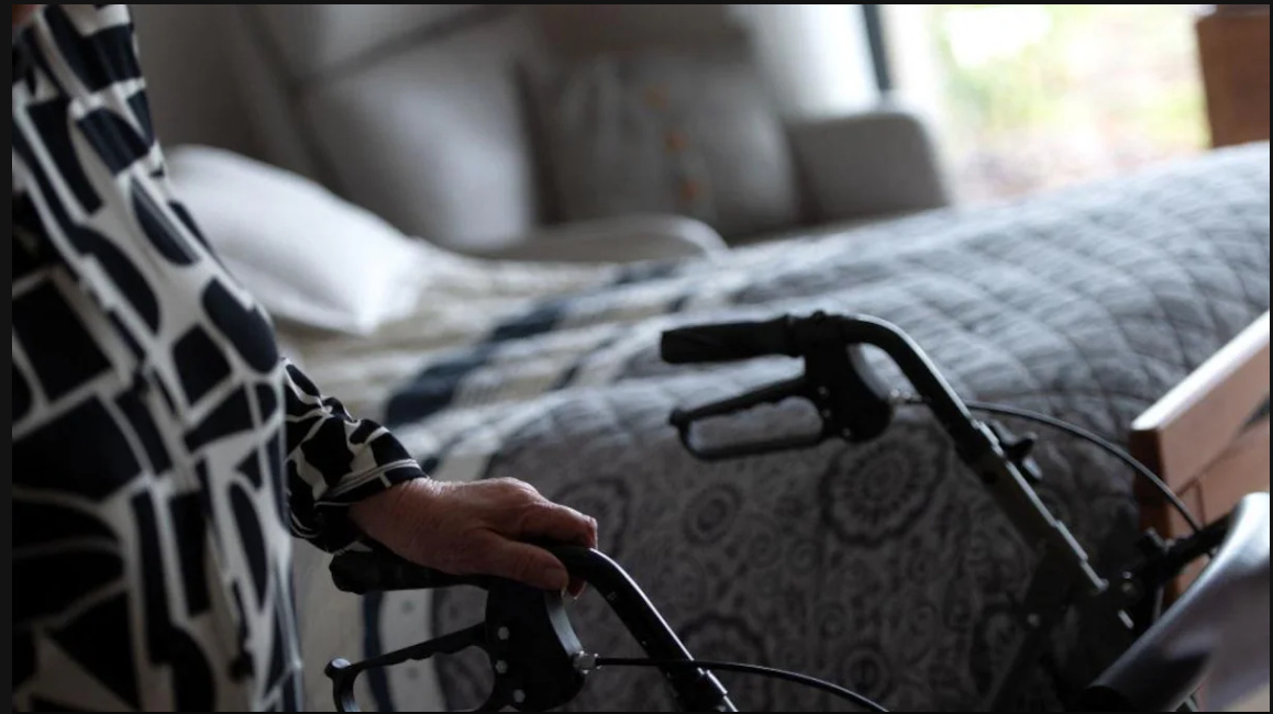 Une personne âgée, dont on voit la main posée sur un déambulateur, à côté d'un lit.