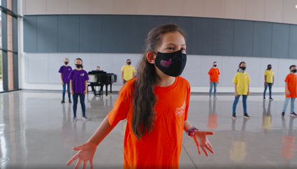 Picture of BC Girls Choir young singer wearing bright orange shirt singing with hands out wide and masked and choir in background in large room and distanced.