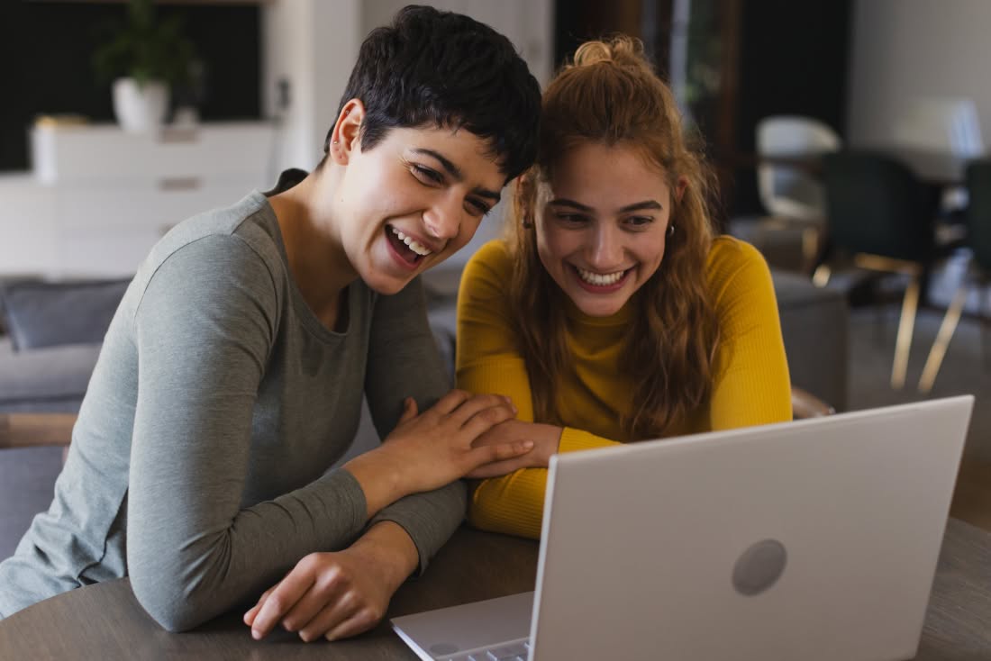 Un couple de femmes souriant consulte un ordinateur portable ensemble, illustrant le soutien communautaire dans leur parcours d'adoption au Québec.