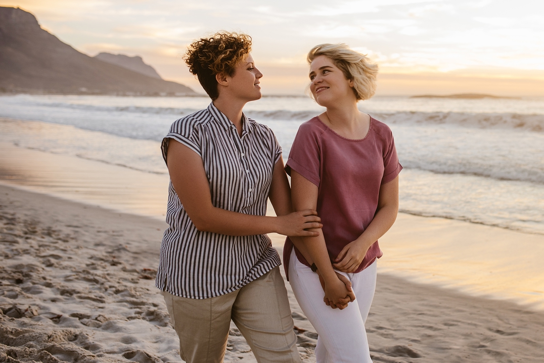 Un couple de femmes se regarde en souriant sur une plage au coucher du soleil, symbole d'engagement dans leur parcours d'adoption au Québec.
