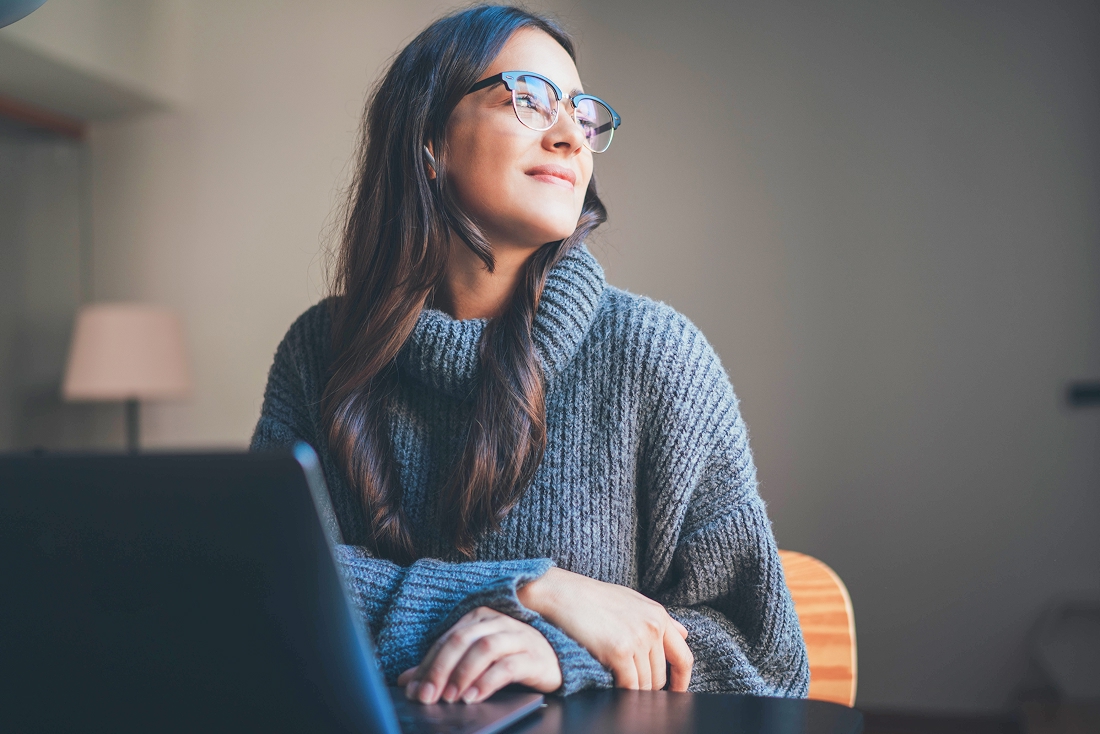 Une femme souriante avec lunettes regarde au loin devant son ordinateur, symbole d'inscription pour devenir membre du parcours d'adoption au Québec.